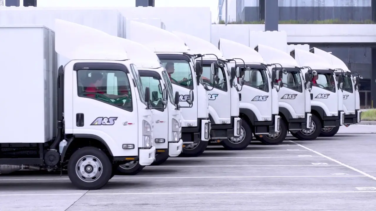A row of white delivery trucks parked in a lot. Each truck is branded with "ASL" on the side. The location appears to be a commercial or industrial area, with buildings in the background and no people visible. The neatly lined-up trucks look ready for a promotional video by a professional video production house.