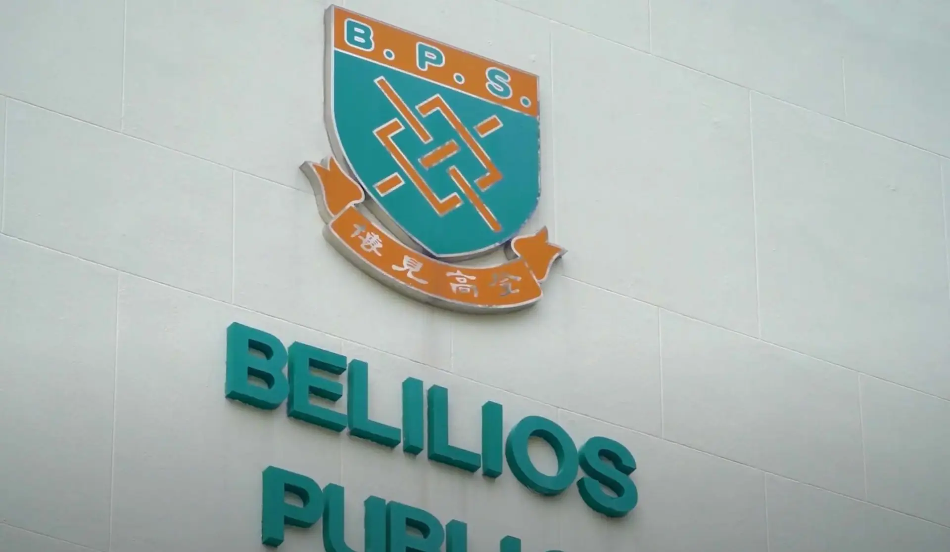 This image showcases the emblem of Belilios Public School on a wall. The emblem features a shield with the initials "B.P.S." and a central design, along with a banner containing Chinese characters. Below the emblem, the school's name is prominently displayed in large teal letters, creating an animation-like visual appeal.