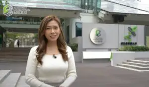 A woman with long wavy hair, wearing a white long-sleeve top, stands outdoors in front of a building with a "Construction Industry Council" sign. The modern building features glass and metal architecture, perfect for an exhibition promotion shoot.