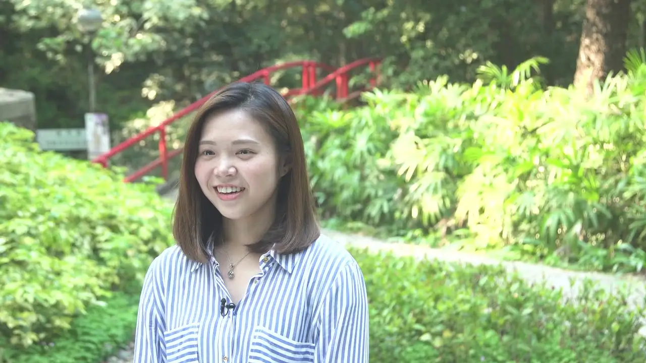 A woman with shoulder-length hair and a striped shirt smiles while standing outdoors. Behind her, there is lush greenery and a red bridge, creating a serene and natural backdrop, perfect for an exhibition promotion video.