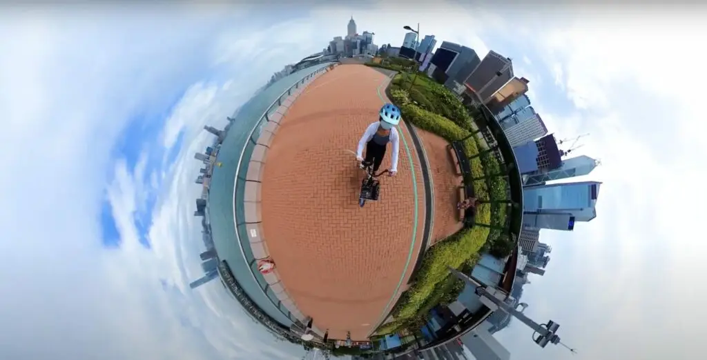A person in a blue helmet is riding a bicycle on a red-brick path near a waterfront. Captured in 360-degree "tiny planet" style, the photo shows city buildings and a cloudy sky encircling the cyclist. This could be part of an innovative promotional video by a top-notch video production house.