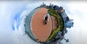 A person in a blue helmet is riding a bicycle on a red-brick path near a waterfront. Captured in 360-degree "tiny planet" style, the photo shows city buildings and a cloudy sky encircling the cyclist. This could be part of an innovative promotional video by a top-notch video production house.