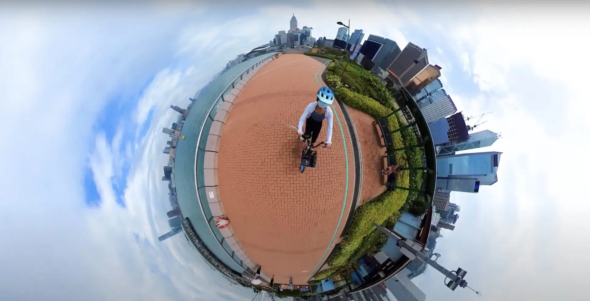 A person in a blue helmet is riding a bicycle on a red-brick path near a waterfront. Captured in 360-degree "tiny planet" style, the photo shows city buildings and a cloudy sky encircling the cyclist. This could be part of an innovative promotional video by a top-notch video production house.