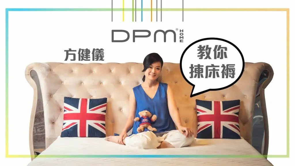 A person sits on a beige tufted sofa with Union Jack pillows on both sides, holding a stuffed animal. The background features colorful lines at the top and Chinese text saying "方健儀" along with a speech bubble that reads "教你撇床褥." The DPM Home logo is displayed above, as part of an exhibition promotion.