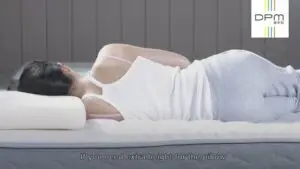 A person lies on their side on a bed, using a white pillow for head support. They are wearing a white tank top and grey pants. Text on the image reads, "If you need extra height for the pillow." The DPM logo features in the upper right corner, making this scene ideal for a promotional video.