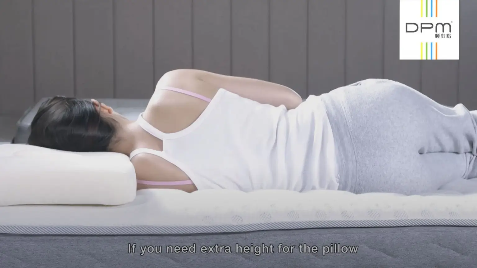 A person lies on their side on a bed, using a white pillow for head support. They are wearing a white tank top and grey pants. Text on the image reads, "If you need extra height for the pillow." The DPM logo features in the upper right corner, making this scene ideal for a promotional video.