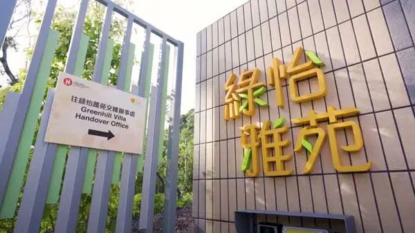 A modern building facade with a sign in both Chinese characters and English reads "Greenhill Villa Handover Office" with an arrow pointing in the direction. The background features greenery and a vertical structure, ideal for video shooting or exhibition promotion.
