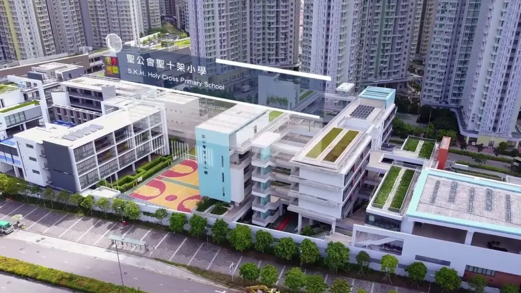 Aerial view of S.K.H. Holy Cross Primary School surrounded by high-rise buildings. The modern school complex features multiple white and light blue buildings, green rooftop spaces, and an outdoor basketball court. A road is visible in the foreground, perfect for a promotional video highlighting the vibrant learning environment.