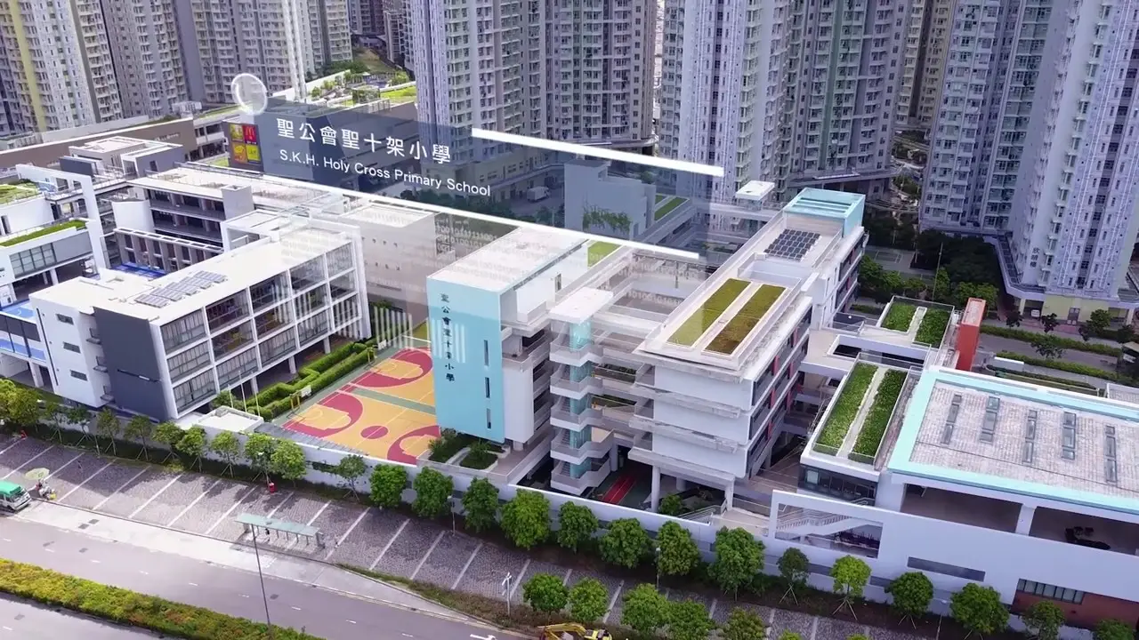 Aerial view of S.K.H. Holy Cross Primary School surrounded by high-rise buildings. The modern school complex features multiple white and light blue buildings, green rooftop spaces, and an outdoor basketball court. A road is visible in the foreground, perfect for a promotional video highlighting the vibrant learning environment.