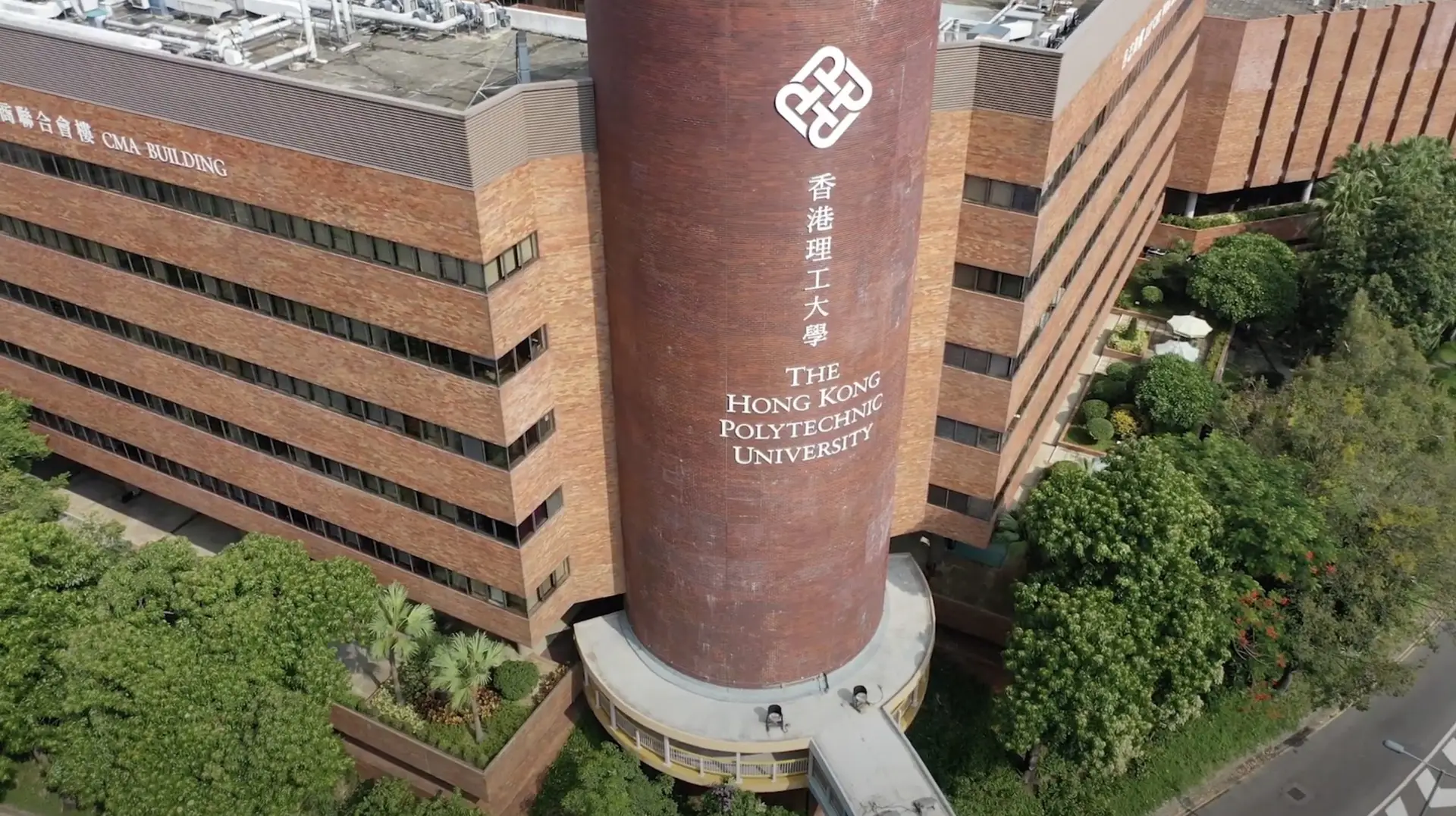 A tall cylindrical building displaying "The Hong Kong Polytechnic University" logo and text is surrounded by other modern brick structures and lush green trees. The aerial view, perfect for animation or video shooting, showcases the university's architectural design and greenery around it.