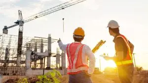 Two construction workers wearing safety gear, including hard hats and reflective vests, stand at a construction site during sunset. One worker is pointing towards a large crane and scaffolding structures, while the other holds a clipboard—capturing the essence of a time-lapse construction video.