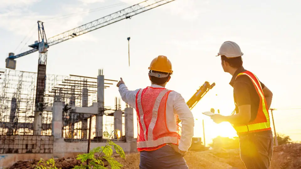 Two construction workers wearing safety gear, including hard hats and reflective vests, stand at a construction site during sunset. One worker is pointing towards a large crane and scaffolding structures, while the other holds a clipboard—capturing the essence of a time-lapse construction video.