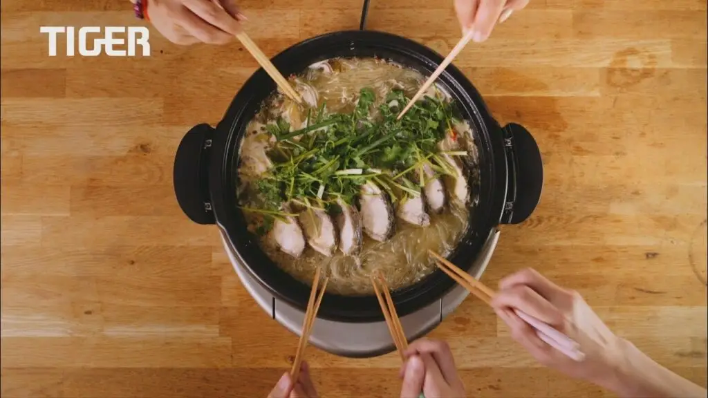 Four pairs of hands holding chopsticks reach into a pot placed on a wooden table. The pot contains broth, noodles, green onions, and slices of meat. Perfect for capturing in an Animation or for a Video Production House to showcase culinary creativity. The word "TIGER" is visible on the left side of the image.