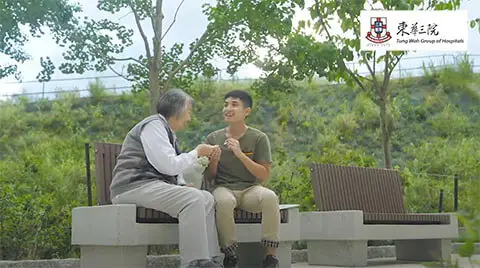 A young man sits on a bench in a park conversing and holding hands with an older woman, possibly his grandmother. Both are smiling. The Tung Wah Group of Hospitals logo is visible in the top right corner, subtly promoting an exhibition. Trees and greenery are in the background.