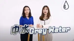 Two women stand next to each other against a plain white background. The woman on the left wears a blue shirt, while the woman on the right dons a white top. The text "Grey Water" appears prominently in front of them with a water drop icon, as part of a promotional video.