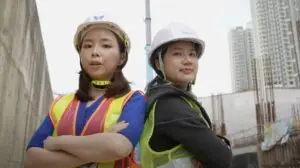 Two construction workers stand back-to-back with arms crossed, wearing hard hats, safety vests, and work attire. They appear confident at an urban construction site with city buildings in the background, ideal for a video production house specializing in construction videos.