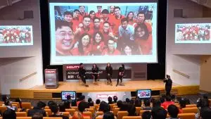 A group of people in red shirts poses for a photo that is projected onto a large screen in a lecture hall. Four people are standing onstage with one person speaking into a microphone. The audience seated in theater-style seats watches the exhibition promotion attentively.