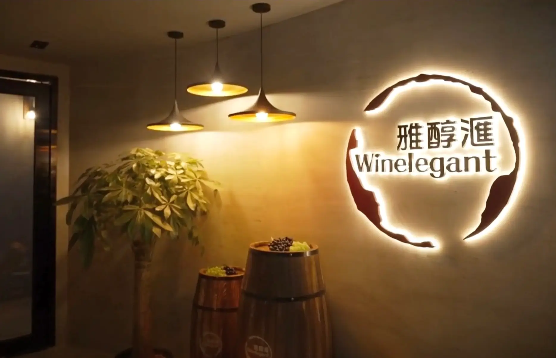 The warmly lit interior of the wine bar features the illuminated "Winelegant" logo on the wall. Three pendant lights hang above two wine barrel tables adorned with grapes, while a lush potted plant adds greenery to the setting, perfect for a promotional video highlighting elegance and charm.