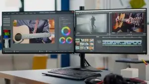 A dual-monitor setup displays a video editing interface. The left screen showcases color correction tools and an image of someone playing a guitar, while the right screen shows a timeline with video clips and an animation of a person dancing. A keyboard and mouse rest on the desk.