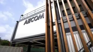 A modern building with a sign that reads "ASCOM" features large vertical wooden beams in the foreground. The facade is white and gray with a partially visible roof deck. The sky is clear with some clouds. The overall appearance, reminiscent of an exhibition promotion, is industrial and sleek.