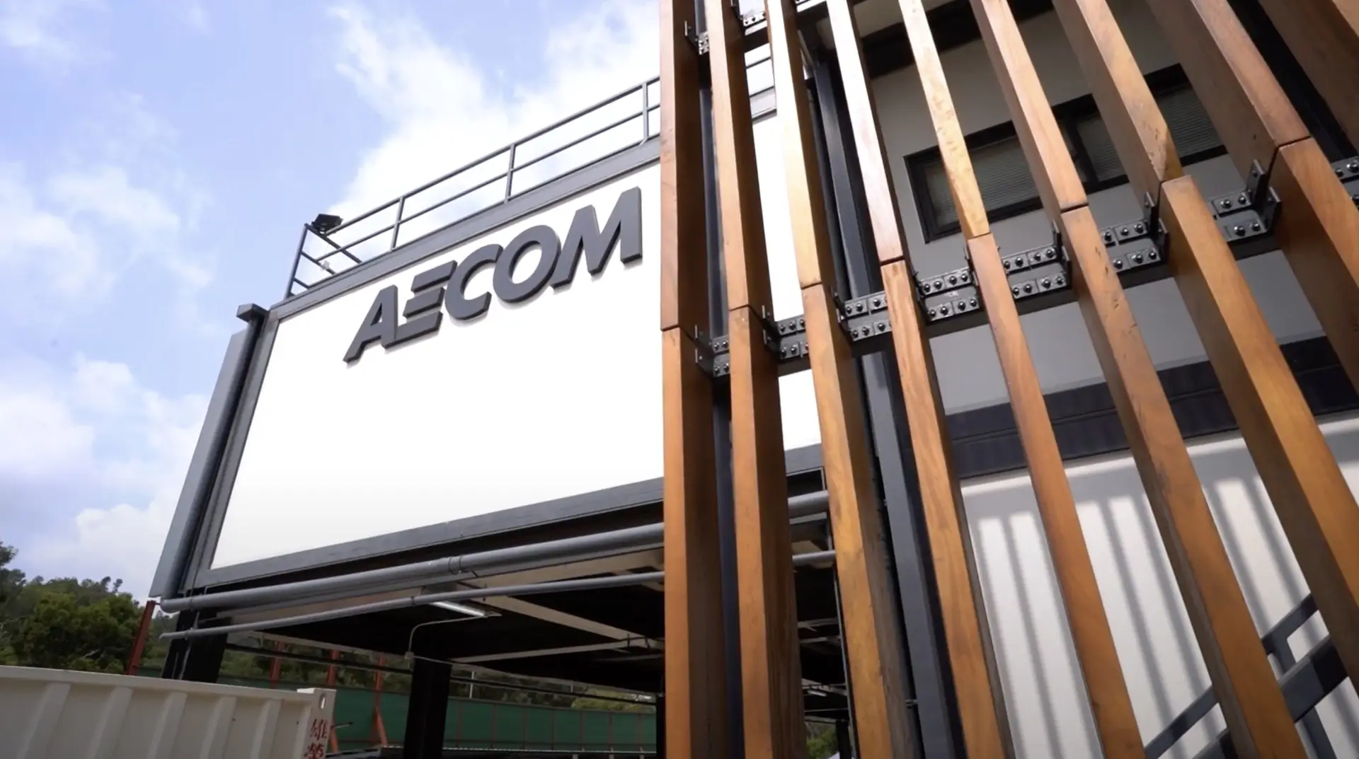 A modern building with a sign that reads "ASCOM" features large vertical wooden beams in the foreground. The facade is white and gray with a partially visible roof deck. The sky is clear with some clouds. The overall appearance, reminiscent of an exhibition promotion, is industrial and sleek.
