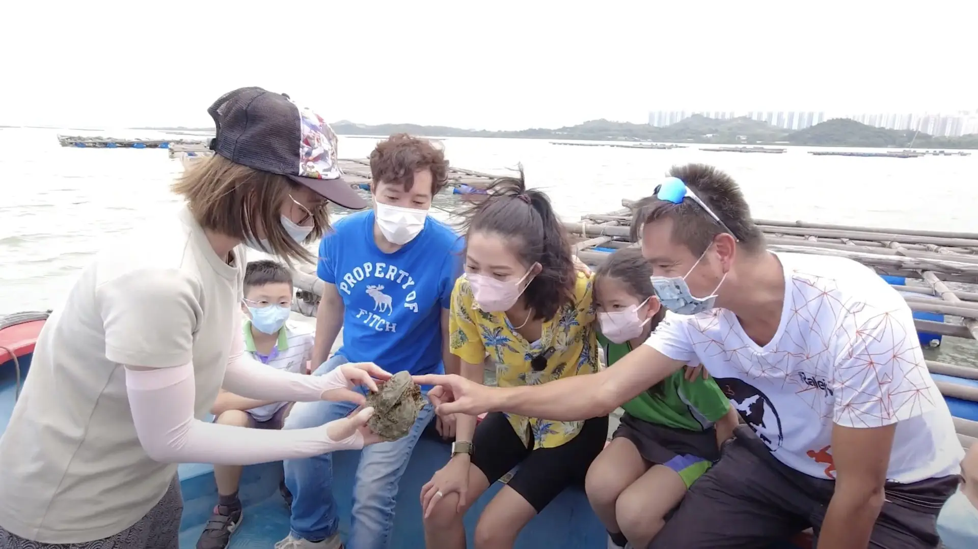 A group of people, all wearing face masks, gather around a person holding a small rock-like object. They are on a boat with the sea and a distant shoreline in the background. Some people, part of a video production house team, reach out to touch the object while others look on curiously.