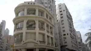 An image showcases a historic three-story building with ornate architectural details, situated at a street corner with high-rise buildings in the background. The building has large rounded windows on the upper floors and a sign with non-Latin script at the top, making it perfect for an exhibition promotion.