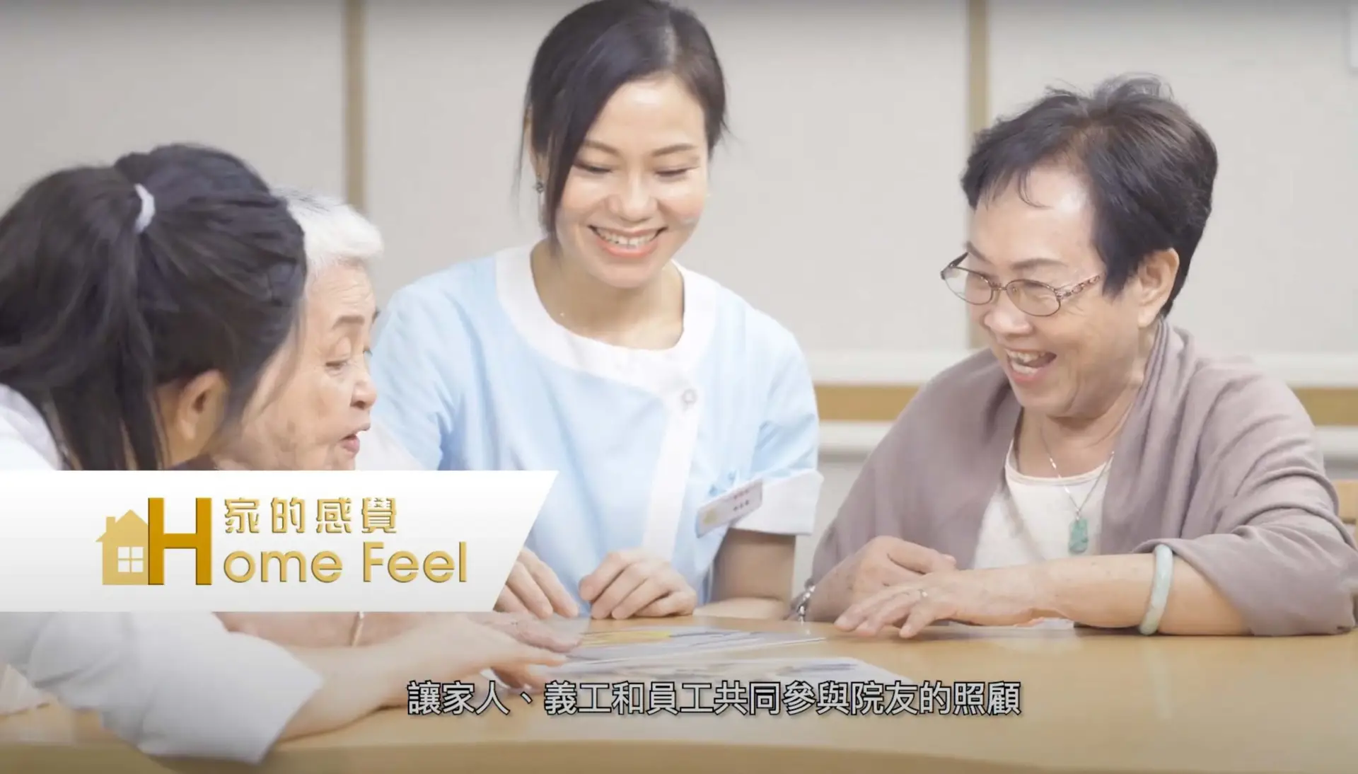 A caregiver and three elderly women are seated around a table, engaging in a warm and lively conversation. They are all smiling and appear joyful, as if part of a promotional video. The text on the image reads "Home Feel" in both English and Chinese.