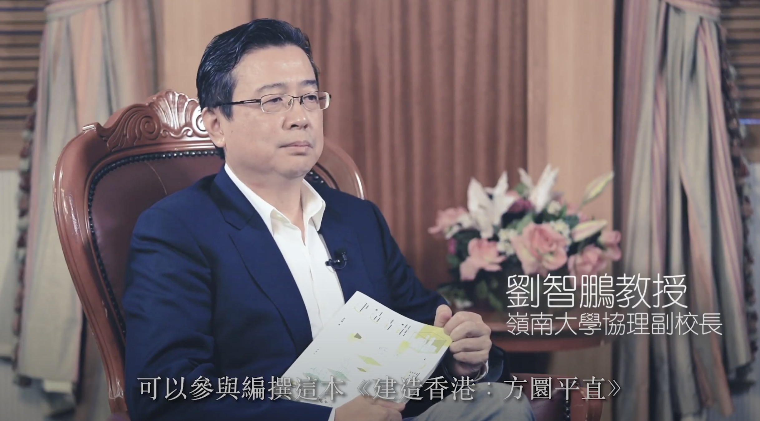 A man in a dark suit and glasses sits in a wooden chair holding a book, with Chinese text on screen. There are pink flowers and beige curtains in the background.