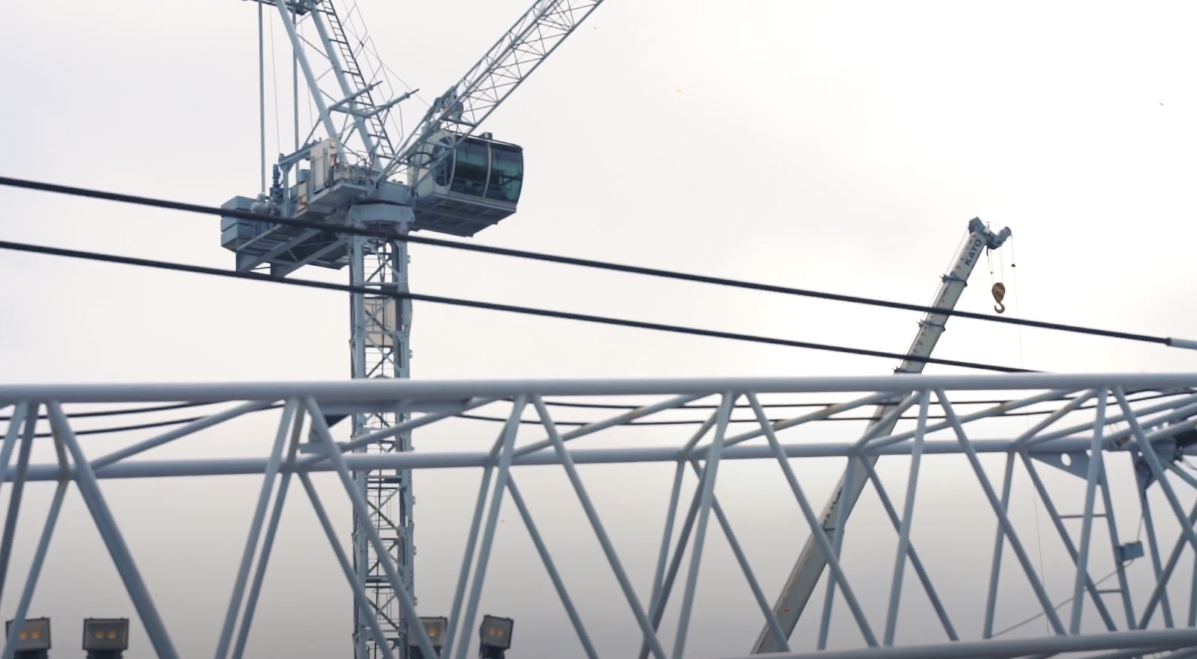 A construction site with two large cranes and a metal framework structure in the foreground against an overcast sky. Power lines cross horizontally through the scene.