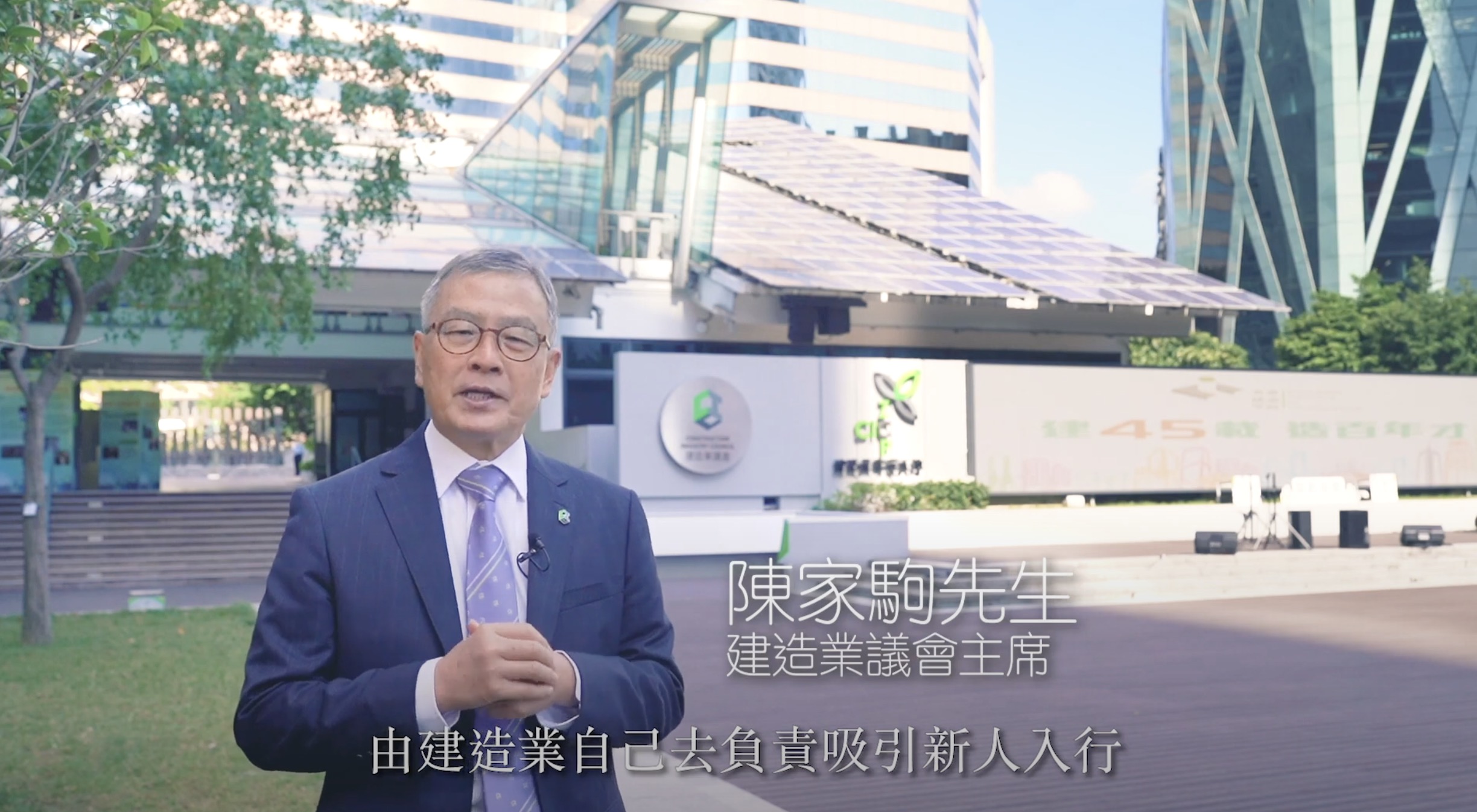 A man in a suit stands outdoors in front of a modern building with glass walls and a solar panel roof, speaking to the camera. Chinese text overlays the image.