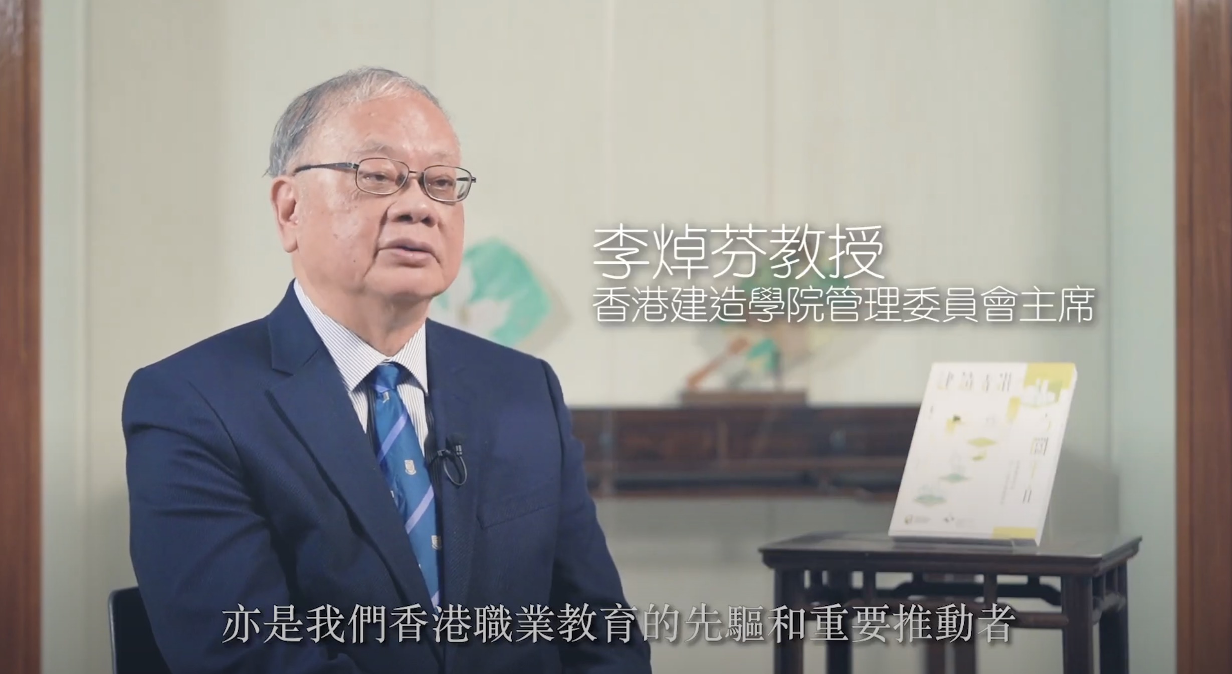 An older man in a suit and glasses sits for an interview. Traditional Chinese art is displayed behind him. Chinese text on screen identifies him as Professor Li Peifang, Chairman of Hong Kong Institute of Construction Management Committee.