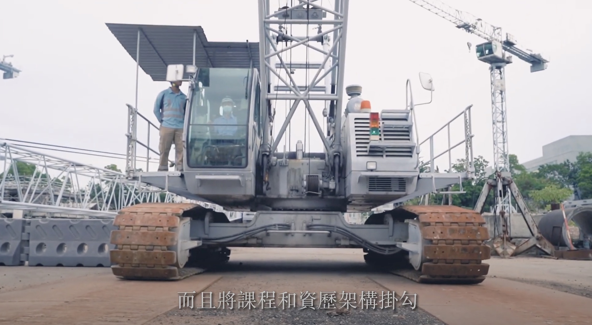 A construction worker stands on a large tracked crane at a building site, with cranes and building materials in the background. Chinese subtitles are displayed at the bottom of the image.