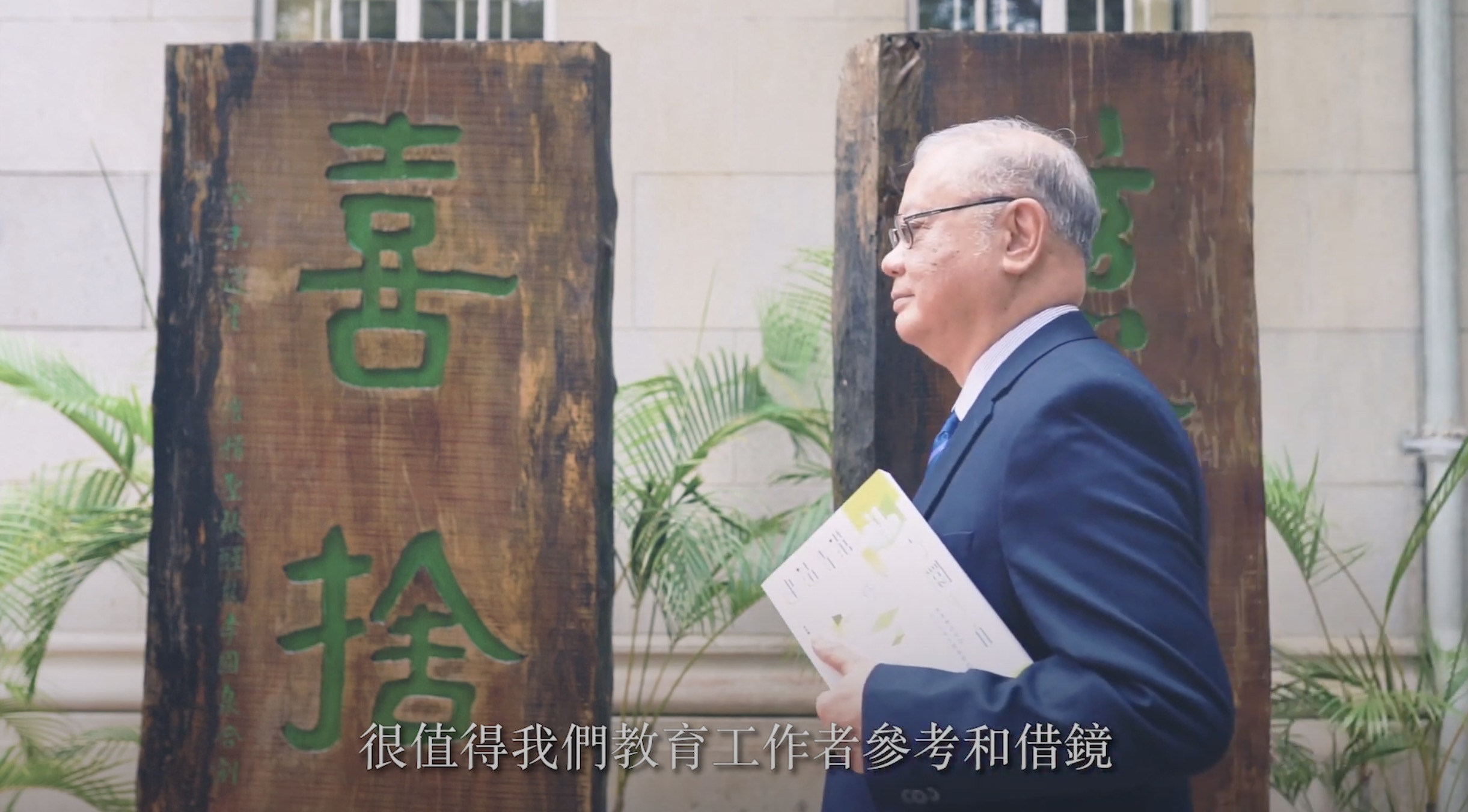 An older man in a suit holds papers and walks outdoors past two large wooden boards with green Chinese characters. Chinese subtitles are visible at the bottom of the image.