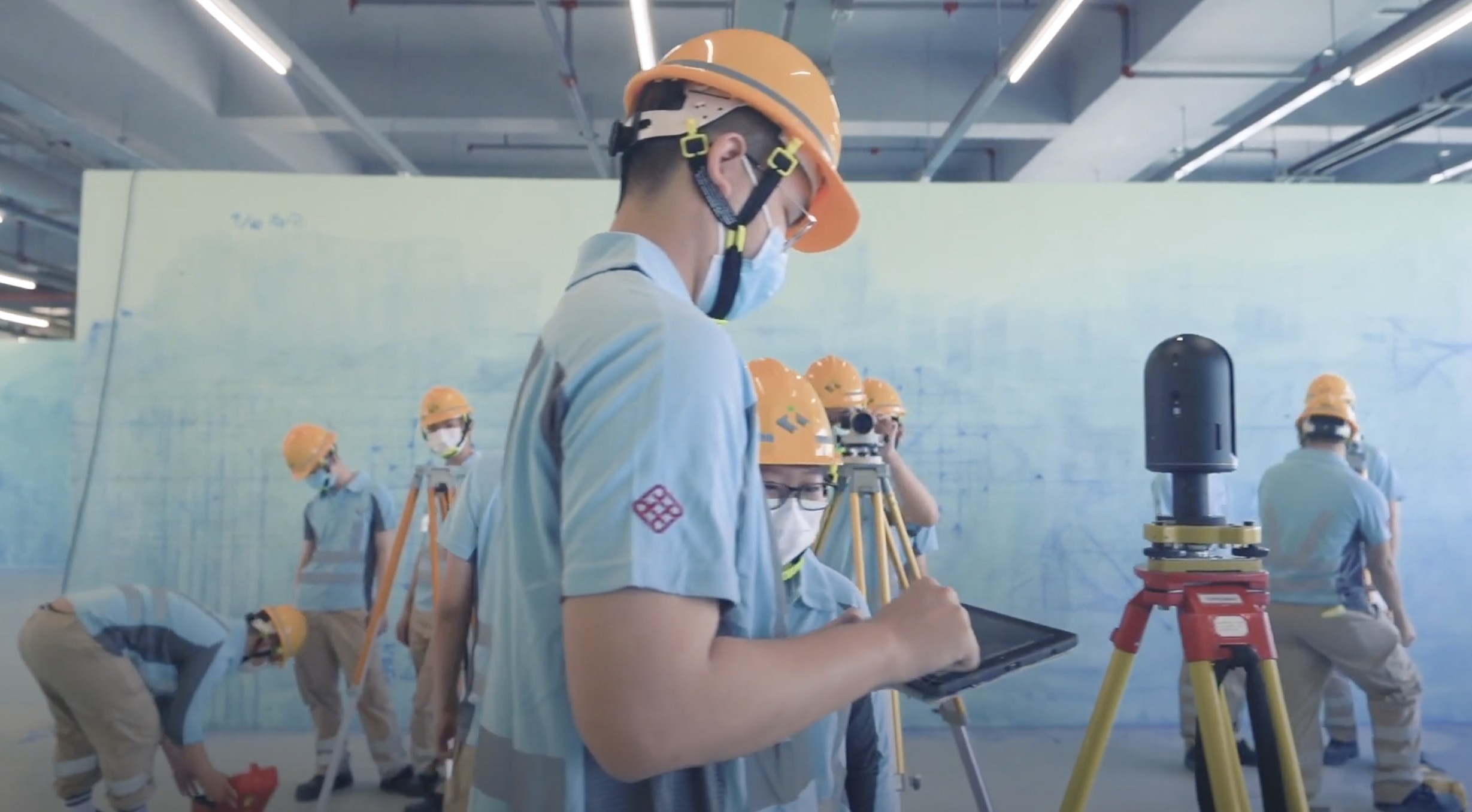 A group of construction workers in blue uniforms and orange helmets work indoors. One worker in the foreground uses a tablet next to survey equipment, while others work in the background. All wear face masks.
