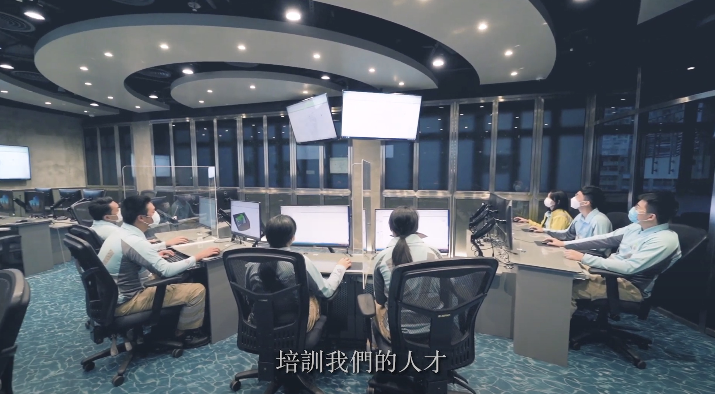 A group of people wearing masks sit at computer workstations in a modern control room. Multiple monitors are mounted in the center. Chinese text appears at the bottom of the image.