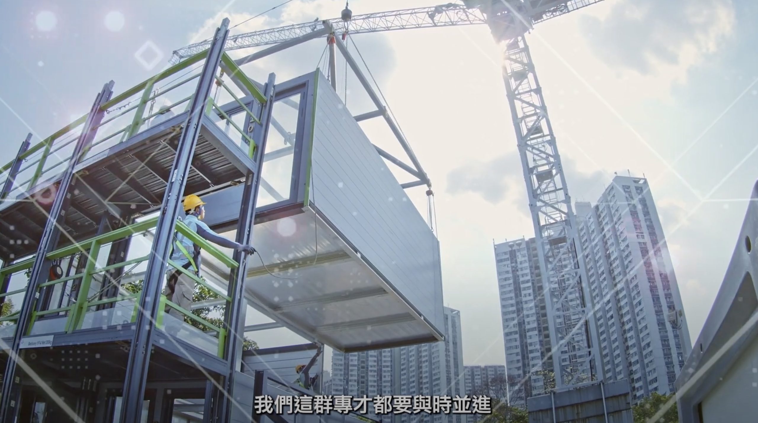 A large crane lifts a modular building unit onto a metal frame at a construction site, with workers in hard hats guiding it. Tall city buildings and another crane are visible in the background. Chinese subtitles appear at the bottom.