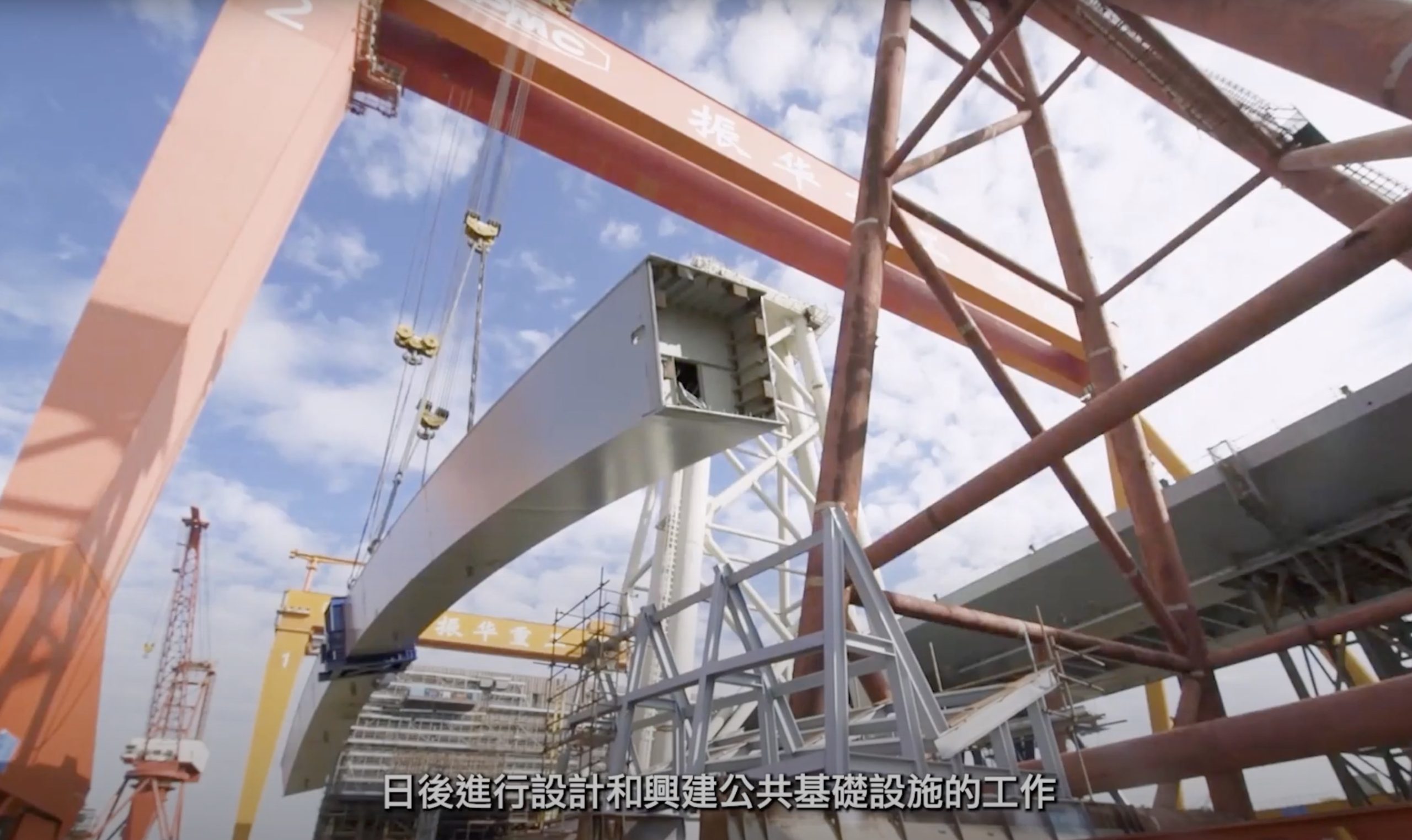 A large steel structure is being assembled at a construction site with cranes around. The sky is clear and there are subtitles in Chinese at the bottom of the image.