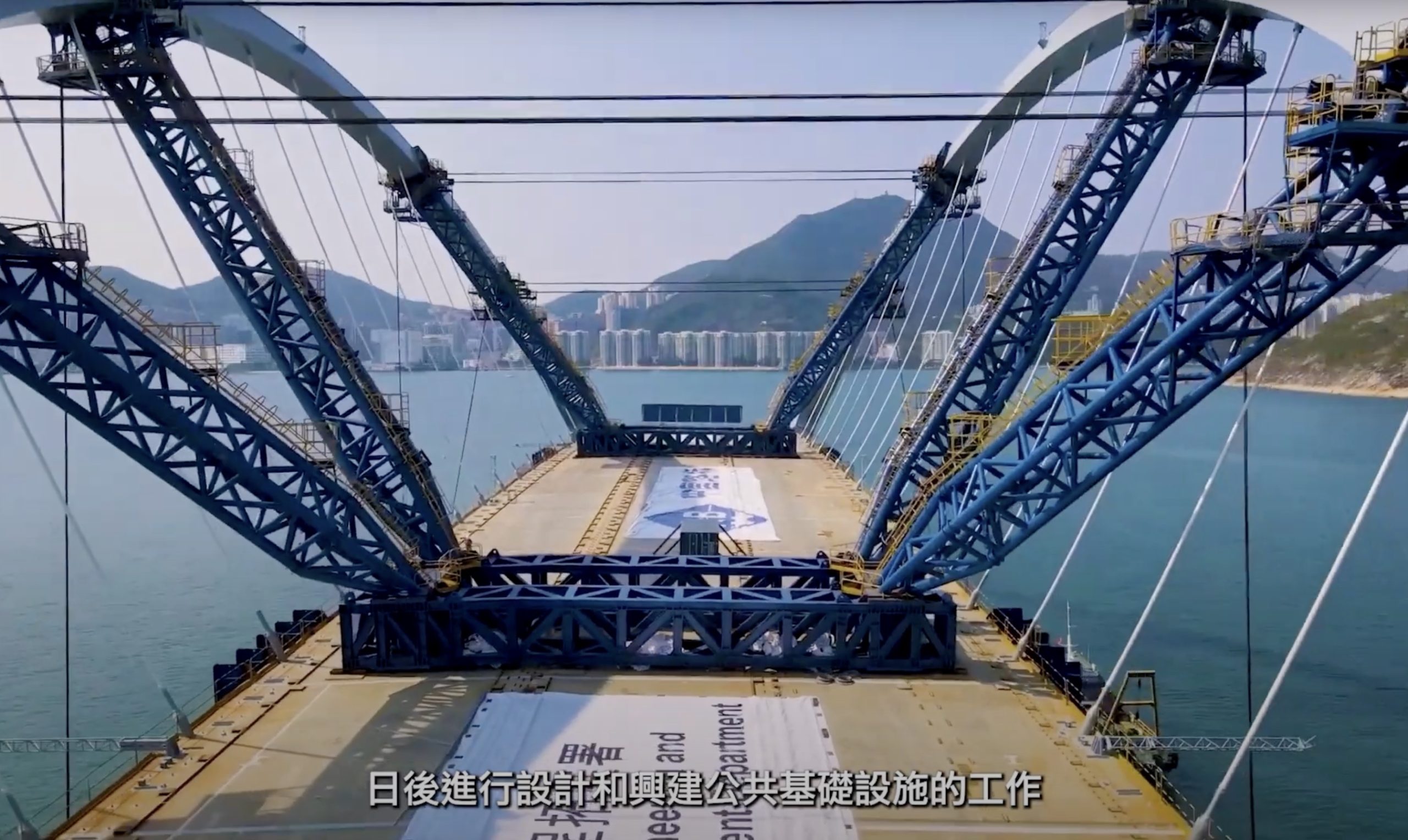 Large blue steel bridge under construction over water, with mountains and a city skyline in the background. Construction materials and signs are visible on the bridge deck. Chinese subtitles appear at the bottom of the image.