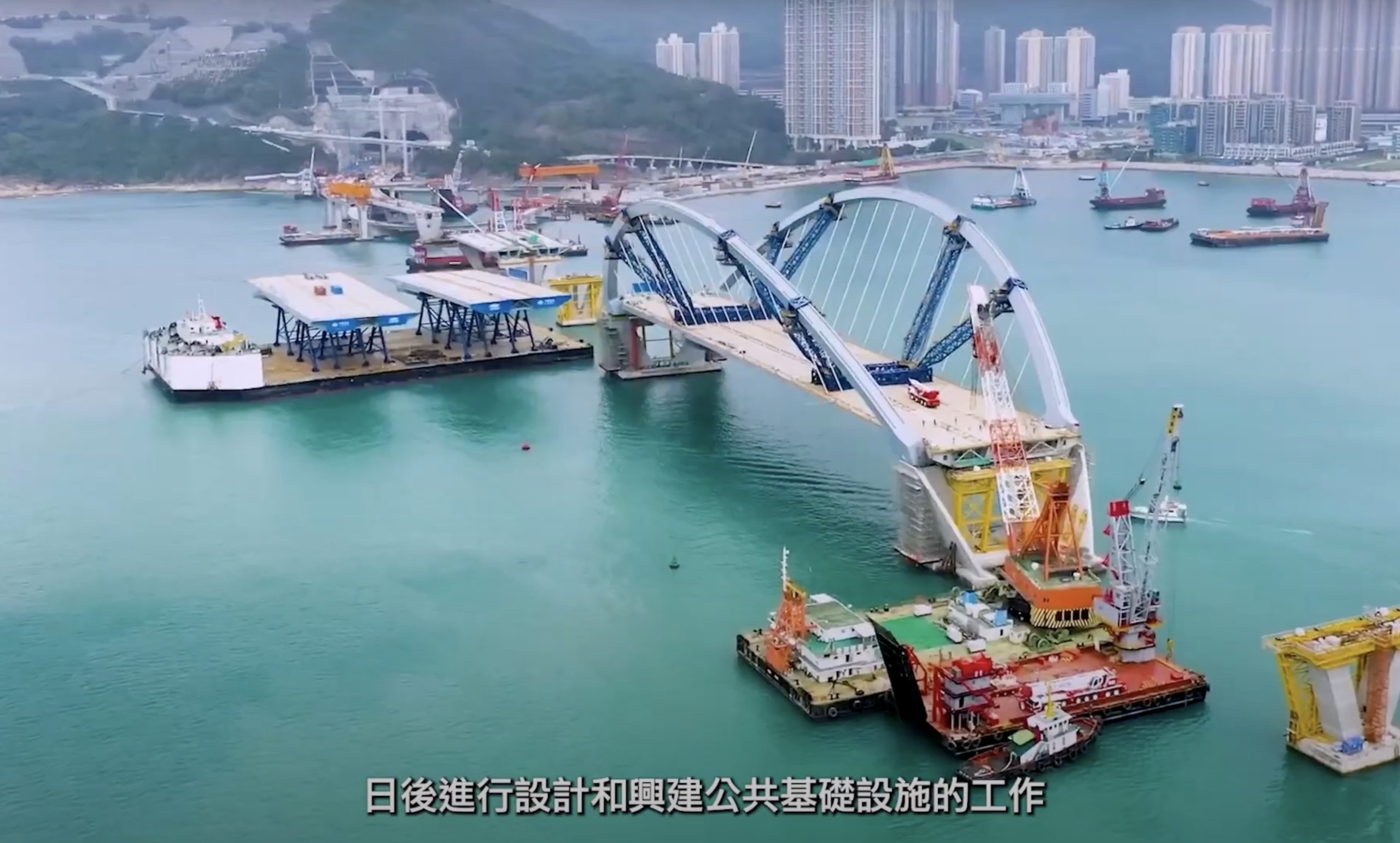 Aerial view of a bridge under construction over water, surrounded by cranes and barges, with a city and hills in the background. Chinese text is displayed at the bottom of the image.