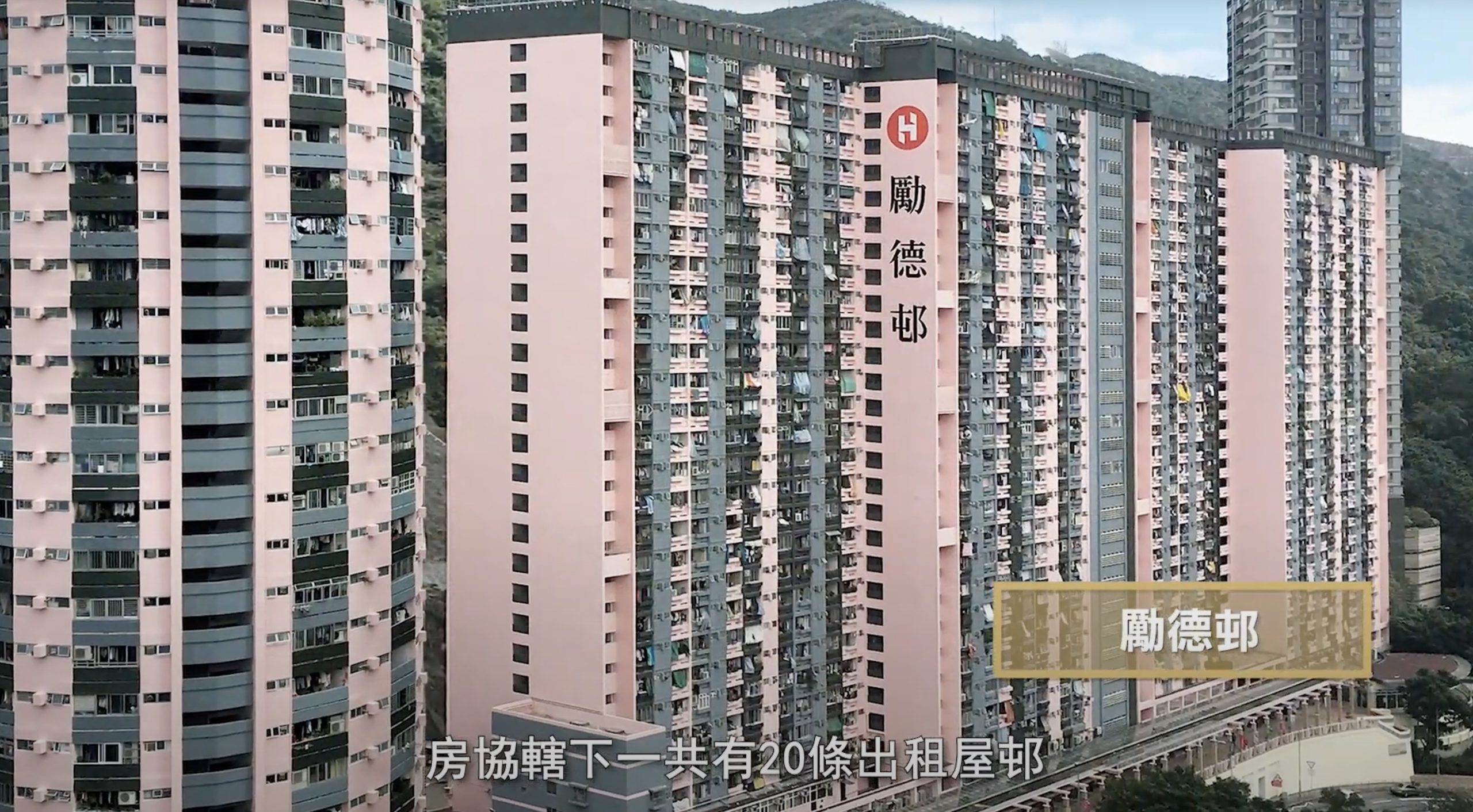 A tall pink and white apartment building with many balconies and windows stands among hills. Large Chinese characters are displayed on the building, and Chinese text appears along the bottom of the image.