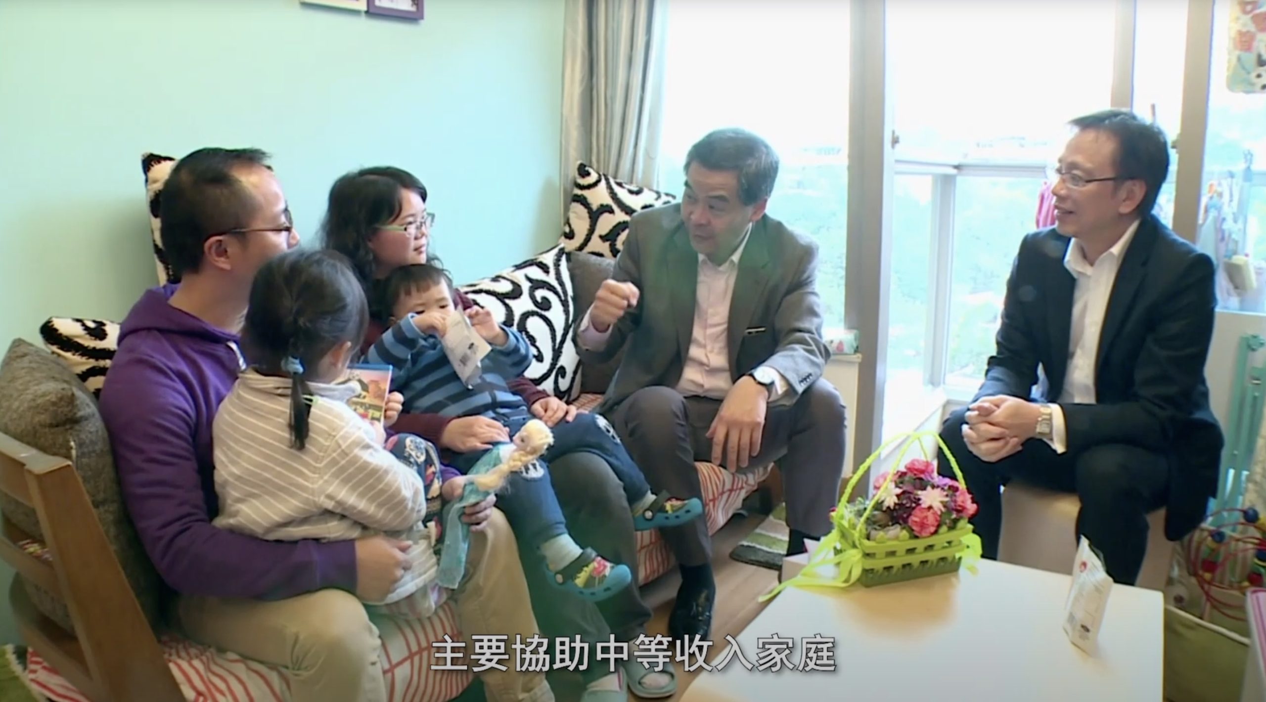 A group of adults and children sit together on a sofa in a living room, engaged in conversation. A flower basket sits on a table nearby. Chinese subtitles appear at the bottom of the image.