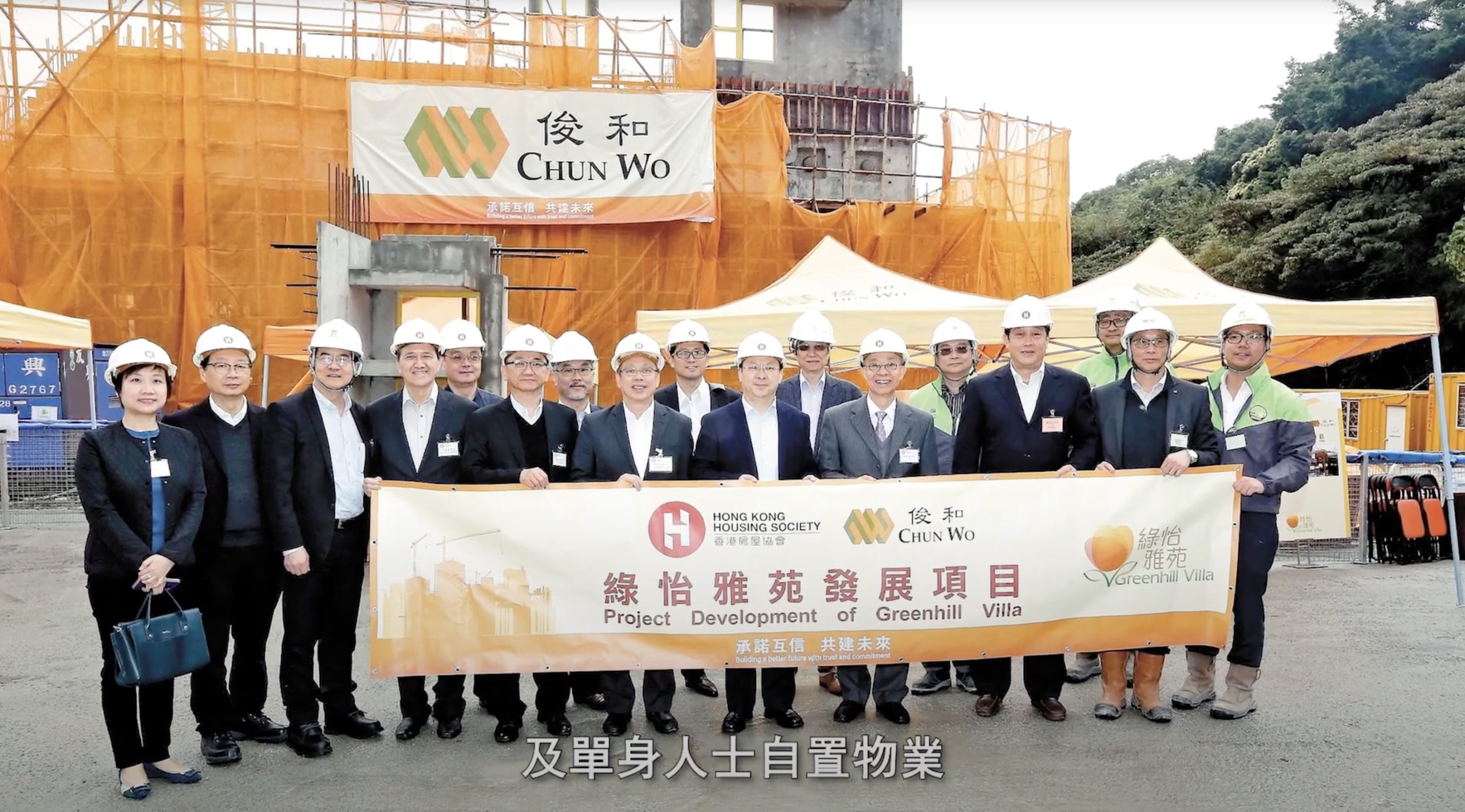 A group of people in construction helmets and suits stand in front of a building site, holding a banner that reads "Project Development of Greenhill Villa." Construction signs and Chinese text are visible in the background.