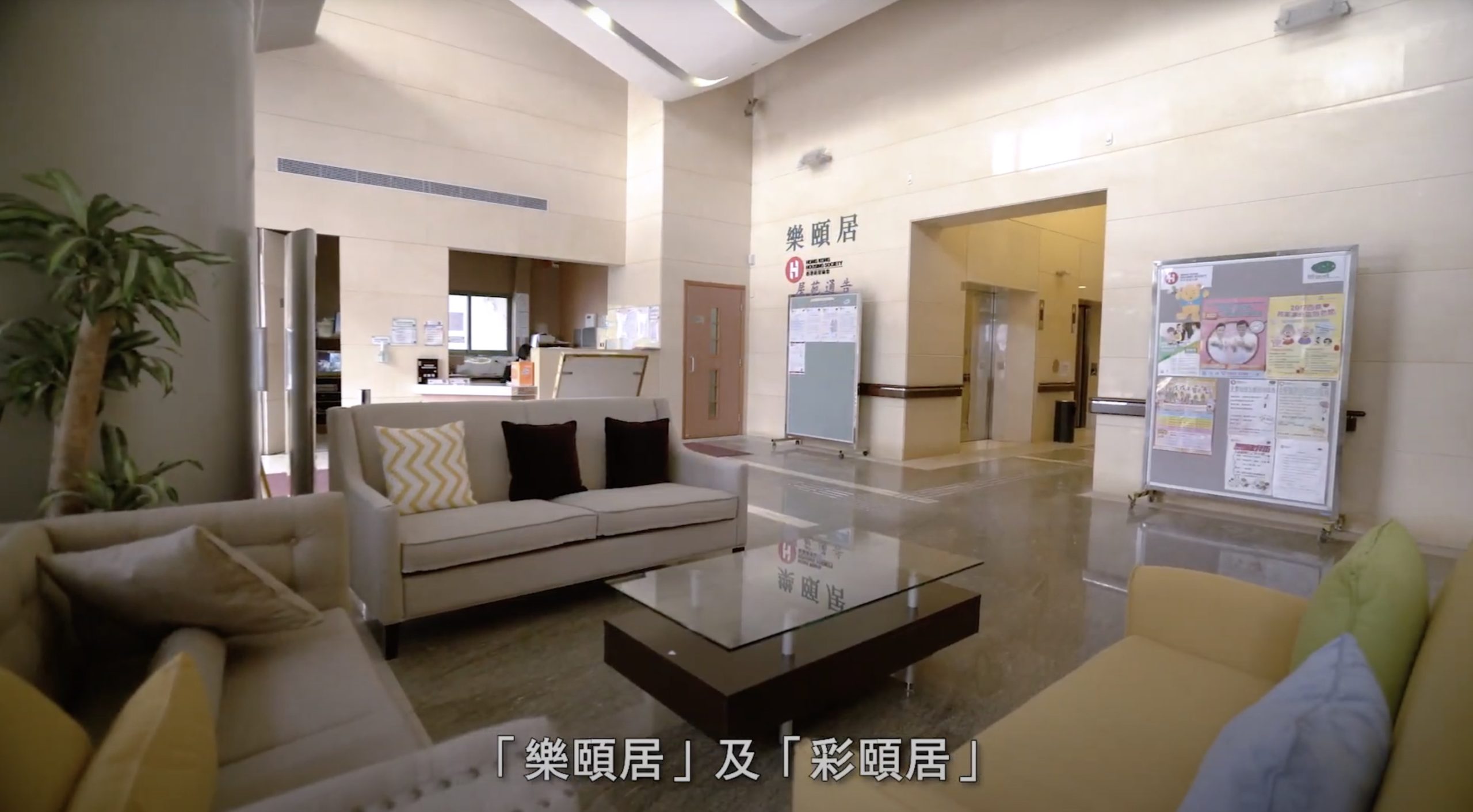 A modern lobby with sofas, a glass coffee table, and bulletin boards on the wall. Chinese text is visible on the wall and as subtitles at the bottom of the image. The space appears bright and welcoming.