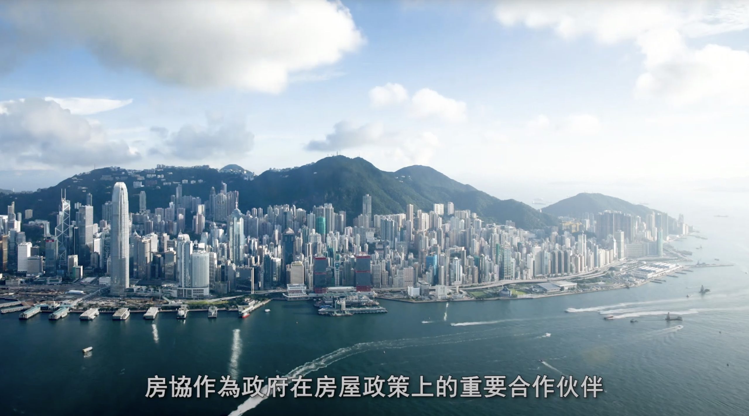 Aerial view of Hong Kong’s skyline with tall skyscrapers along the waterfront and green mountains in the background. Chinese subtitles appear at the bottom of the image.