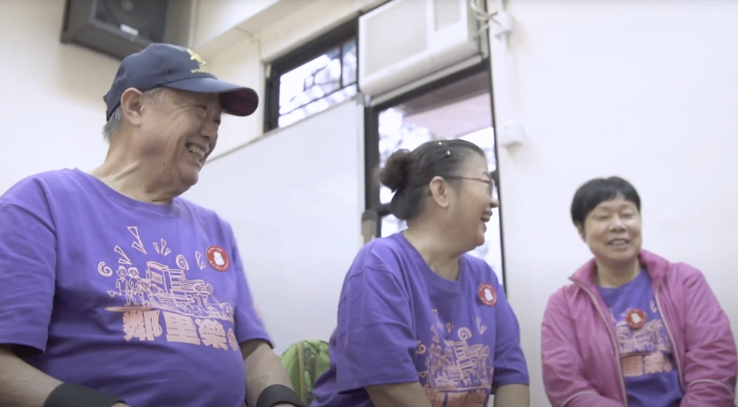 Three older adults sit together indoors, smiling and laughing. They all wear matching purple shirts with a graphic and red buttons, and one person wears a blue cap. The atmosphere appears friendly and joyful.