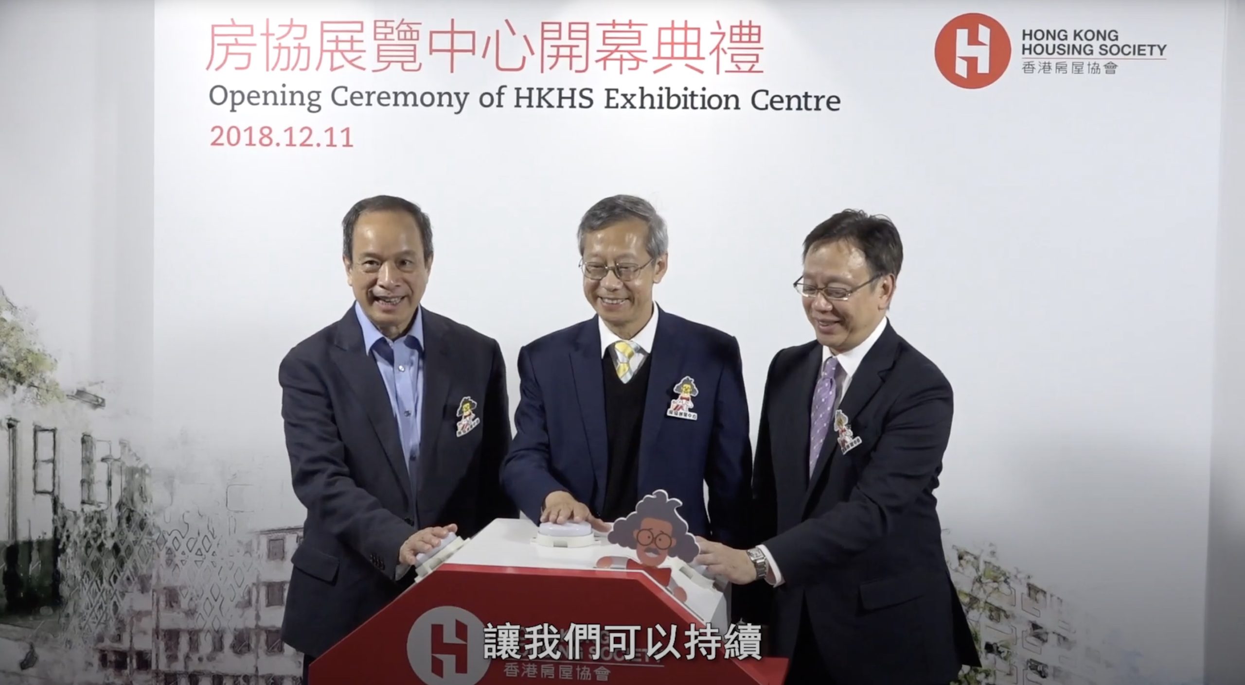 Three men in suits stand together, smiling, with their hands on a podium at the Opening Ceremony of the HKHS Exhibition Centre on December 11, 2018. The Hong Kong Housing Society logo is displayed in the background.