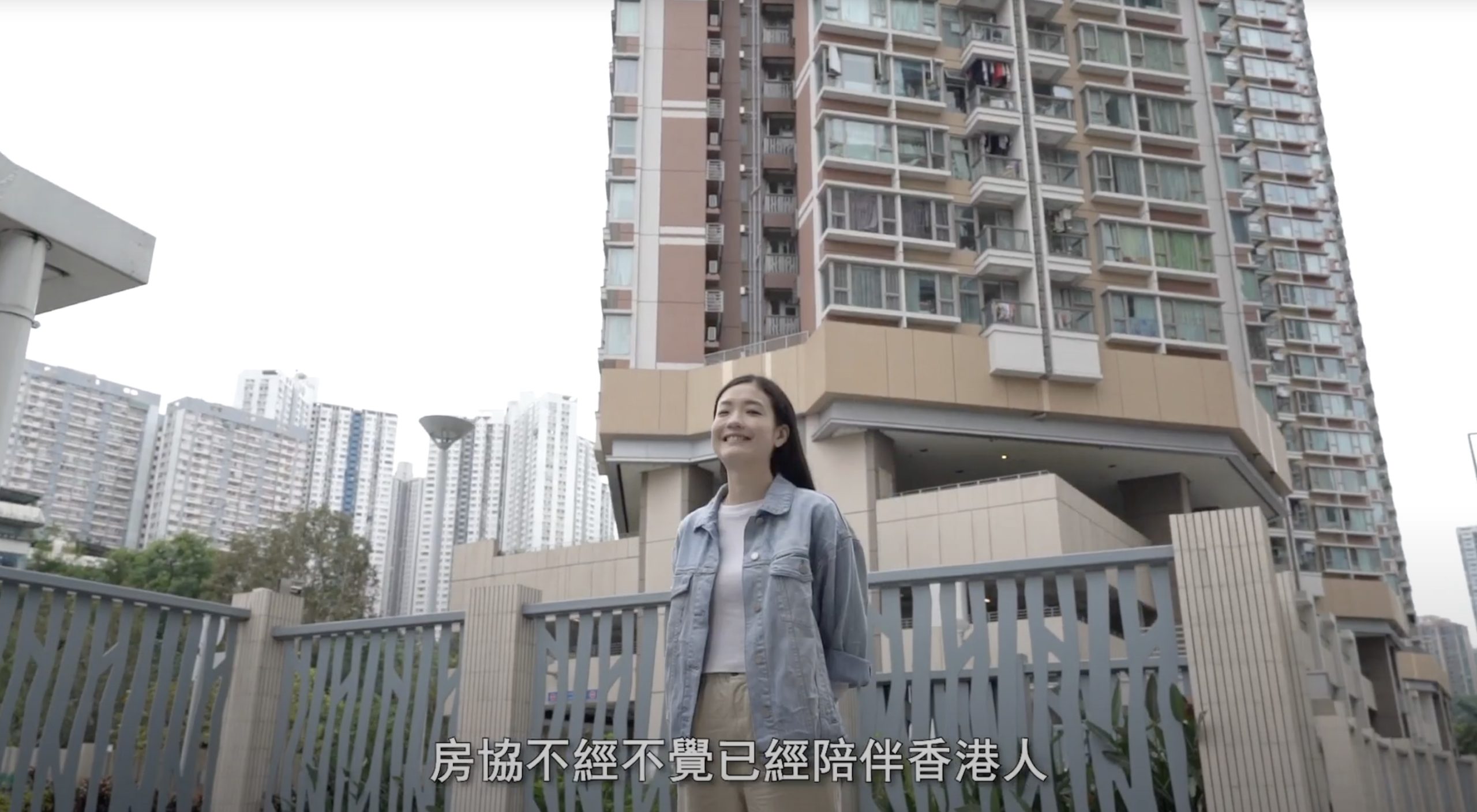 A woman in a denim jacket stands and smiles outside a high-rise residential building, with more tall buildings in the background. Chinese text appears at the bottom of the image.