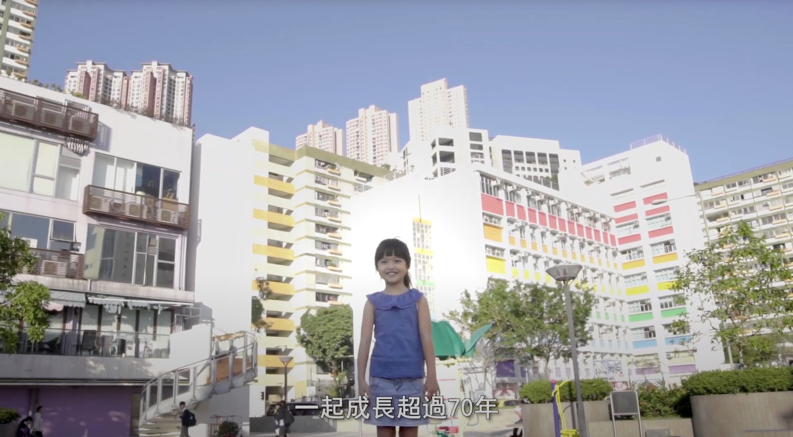 A young girl in a blue dress stands smiling in front of colorful apartment buildings on a sunny day. Chinese text appears at the bottom of the image. Tall skyscrapers are visible in the background.
