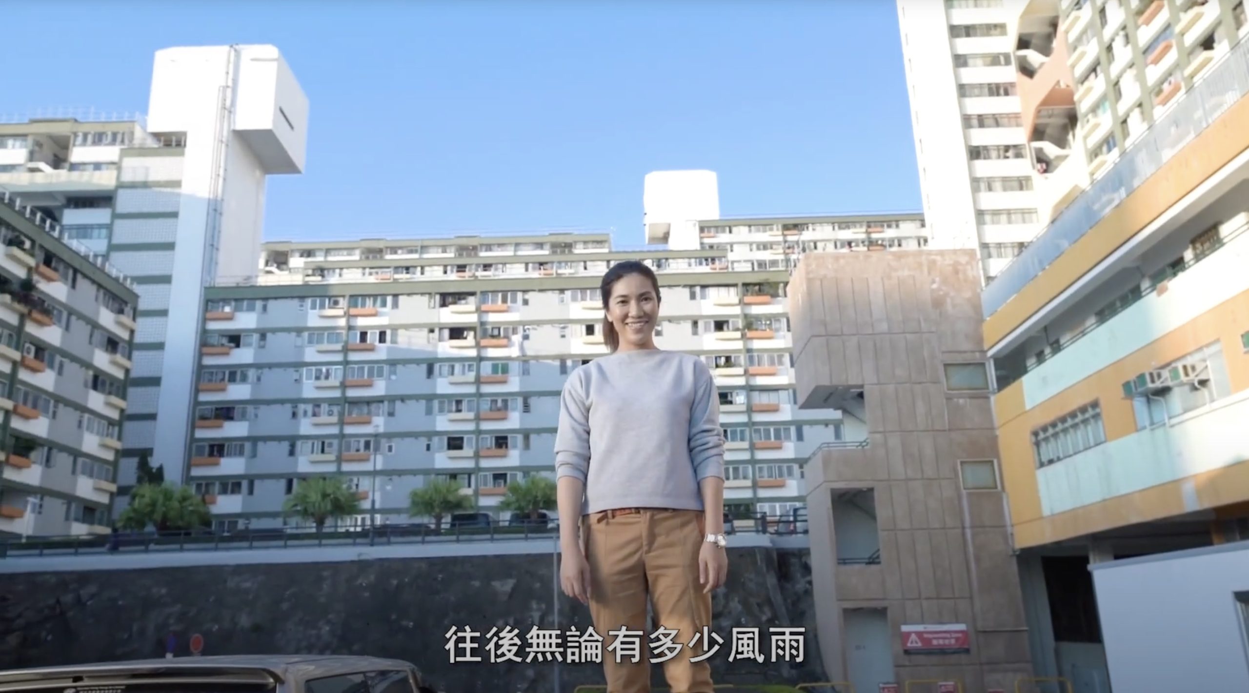 A woman smiles while standing in a parking lot, with tall residential buildings behind her under a clear sky. Chinese subtitles appear at the bottom of the image.